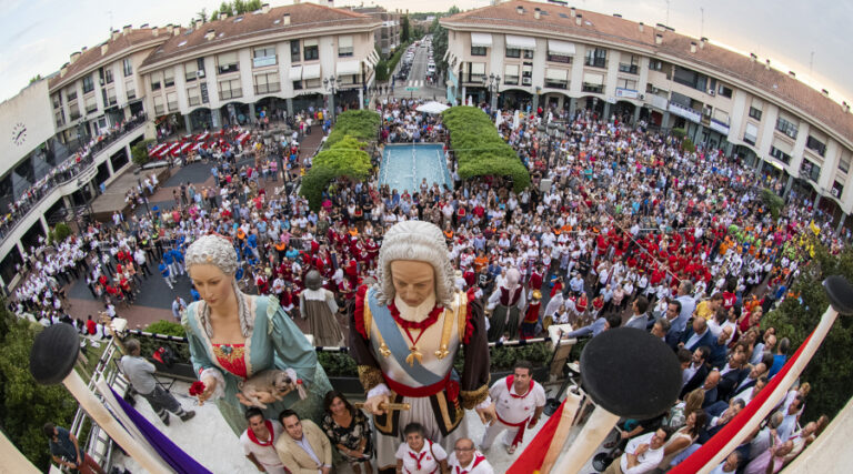 Vista aérea de una plaza repleta de público con gigantes y cabezudos durante el pregón de las fiestas patronales.